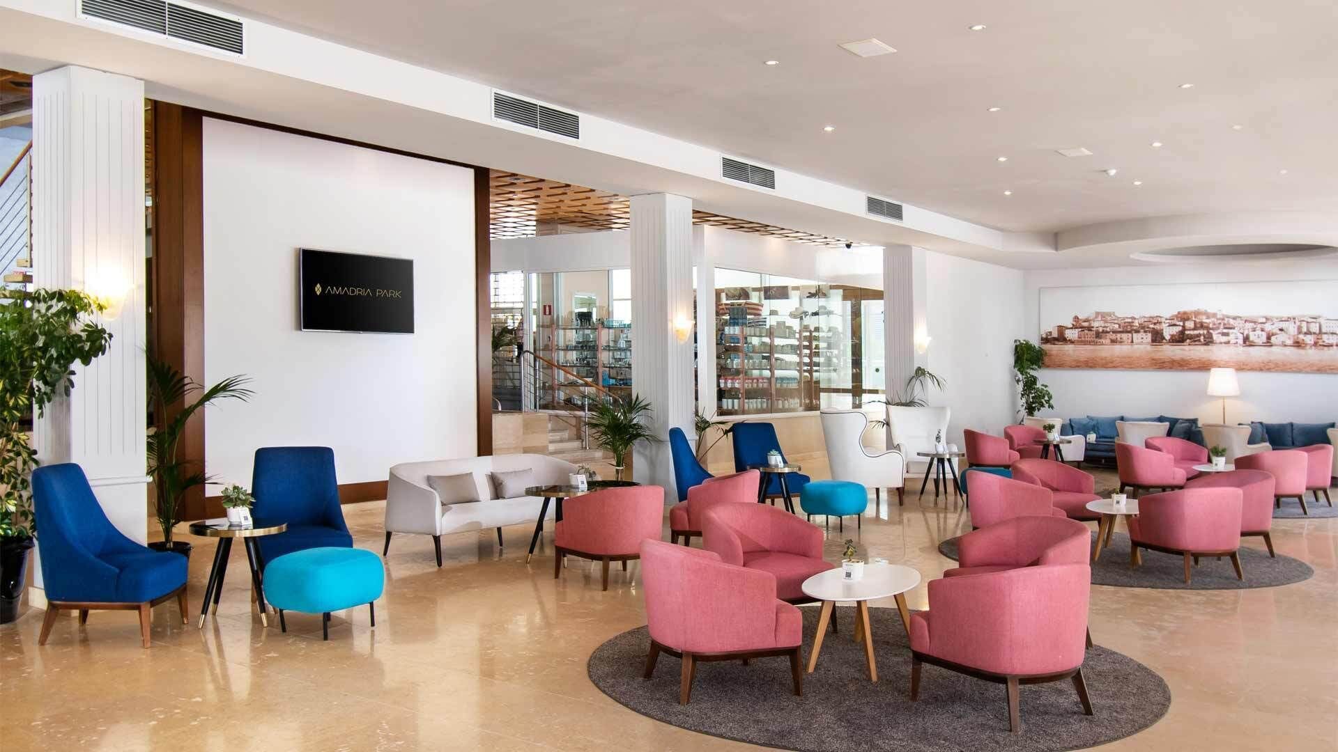 A spacious lobby with a variety of seating options, including blue and pink chairs, a sofa, and a coffee table. There is a large TV screen displaying 'AMADRIA PARK' and a bookshelf filled with books.