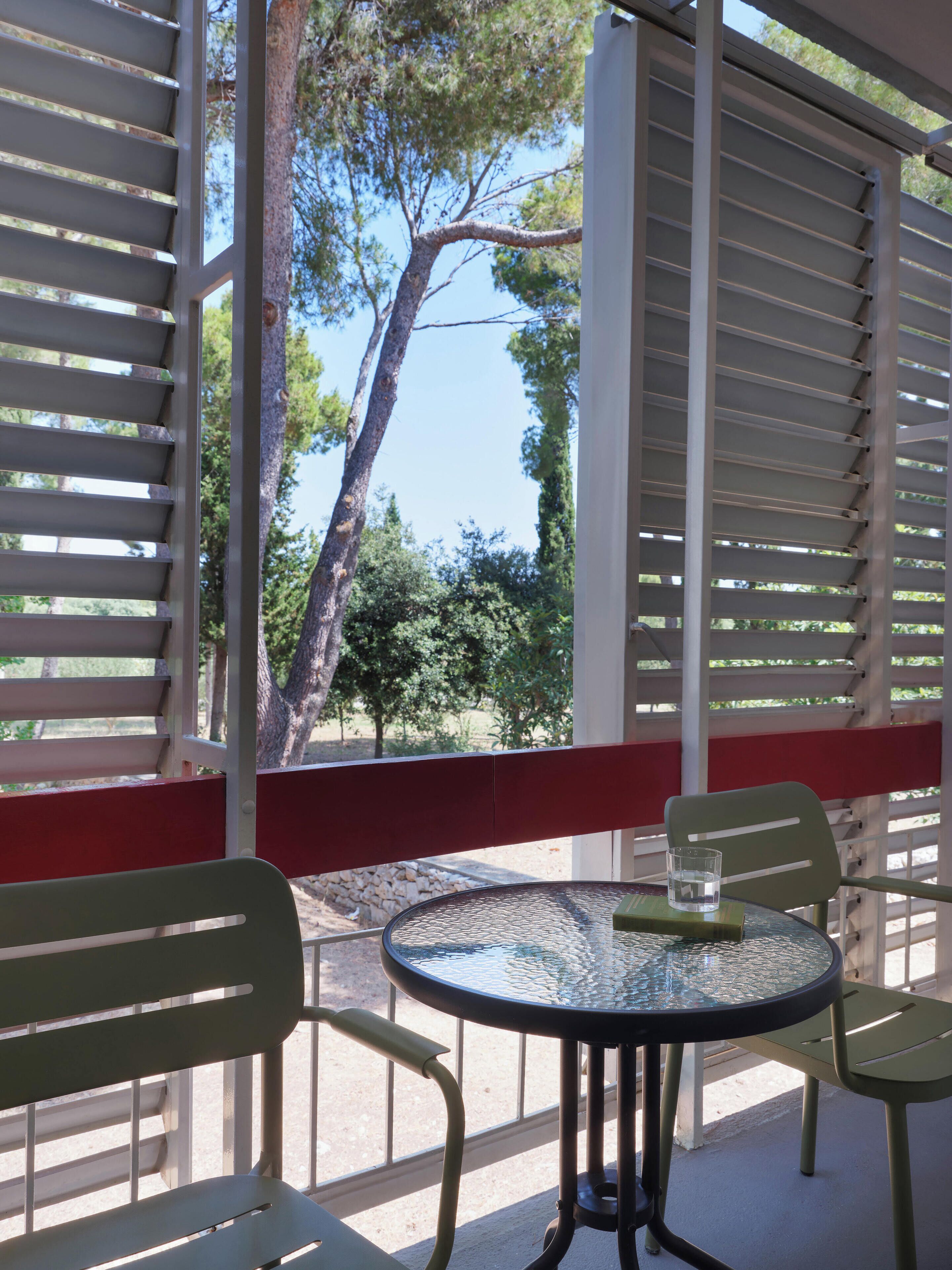 A covered outdoor seating area with a table and chairs, a glass top, and a view of trees and sky through open white blinds.
