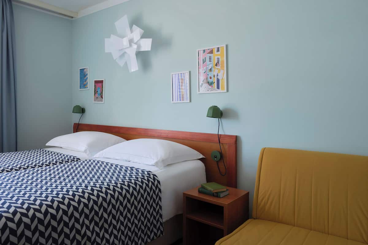 A hotel room with a bed, a nightstand, a lamp, a chair, and wall art.