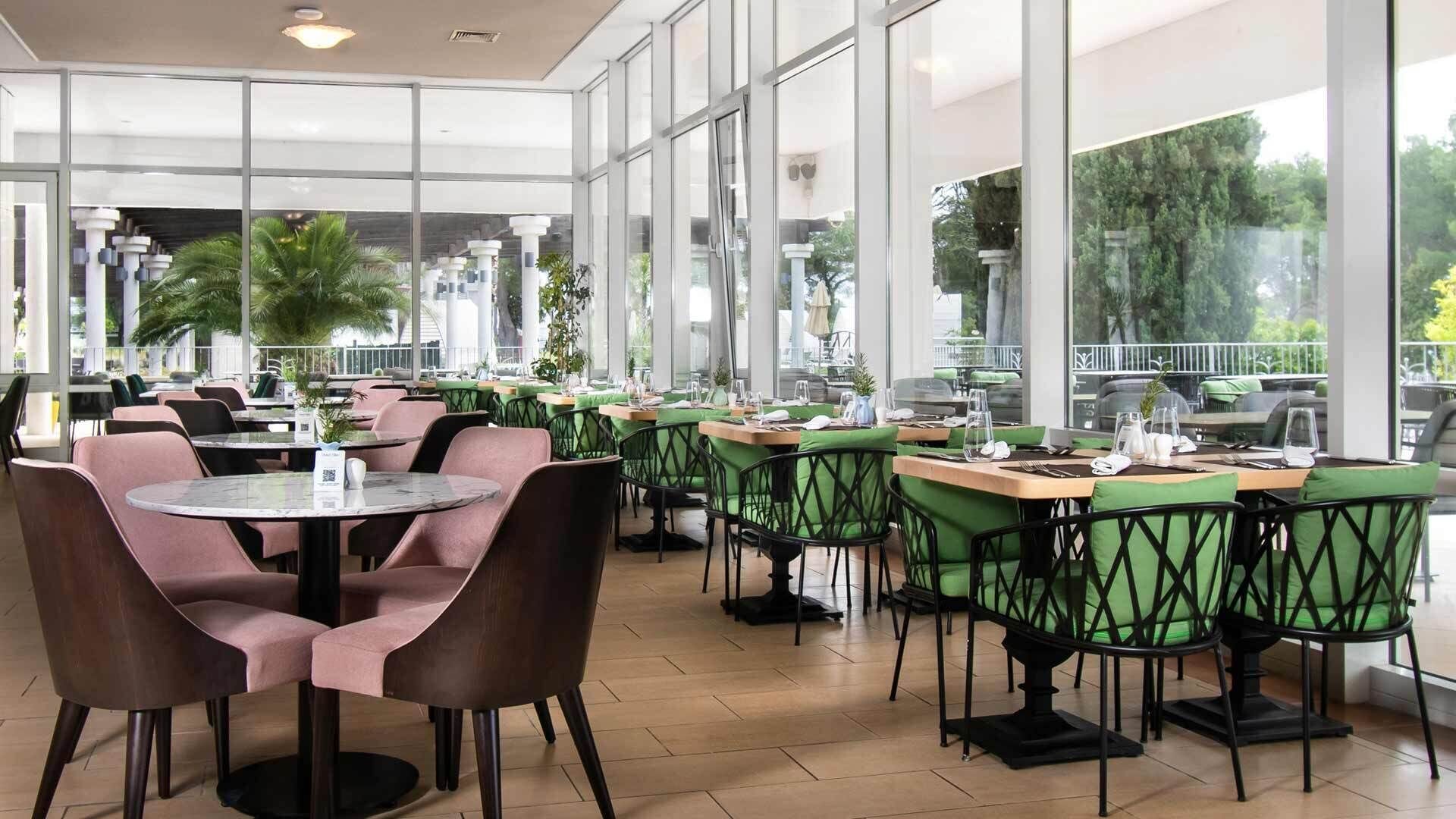 Ein Restaurant mit runden Tischen und grünen Stühlen, großen Fenstern und Zimmerpflanzen.