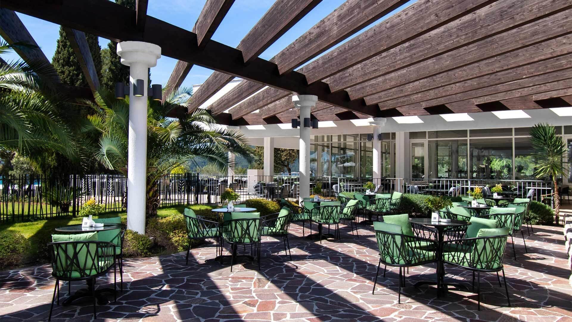 An outdoor patio area with stone flooring, green metal chairs, and tables, surrounded by a wooden pergola structure.