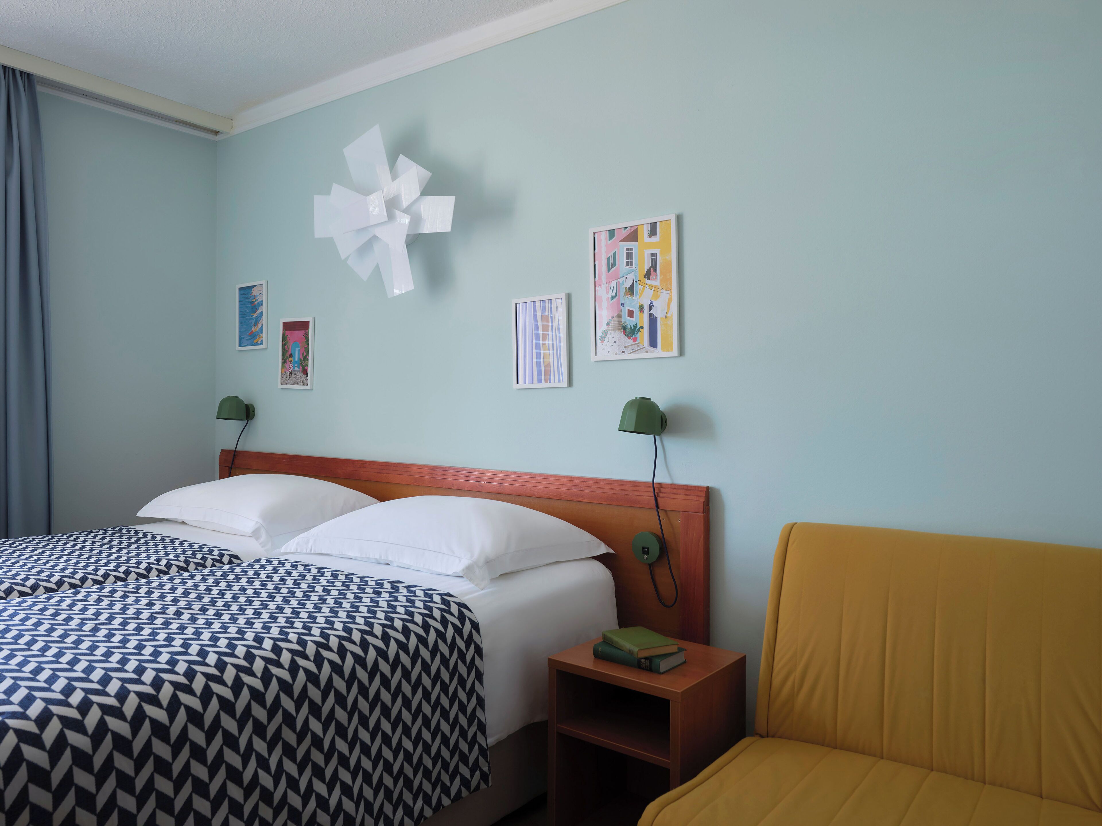 Une chambre d’hôtel avec un lit, une table de chevet, une lampe et un canapé jaune.