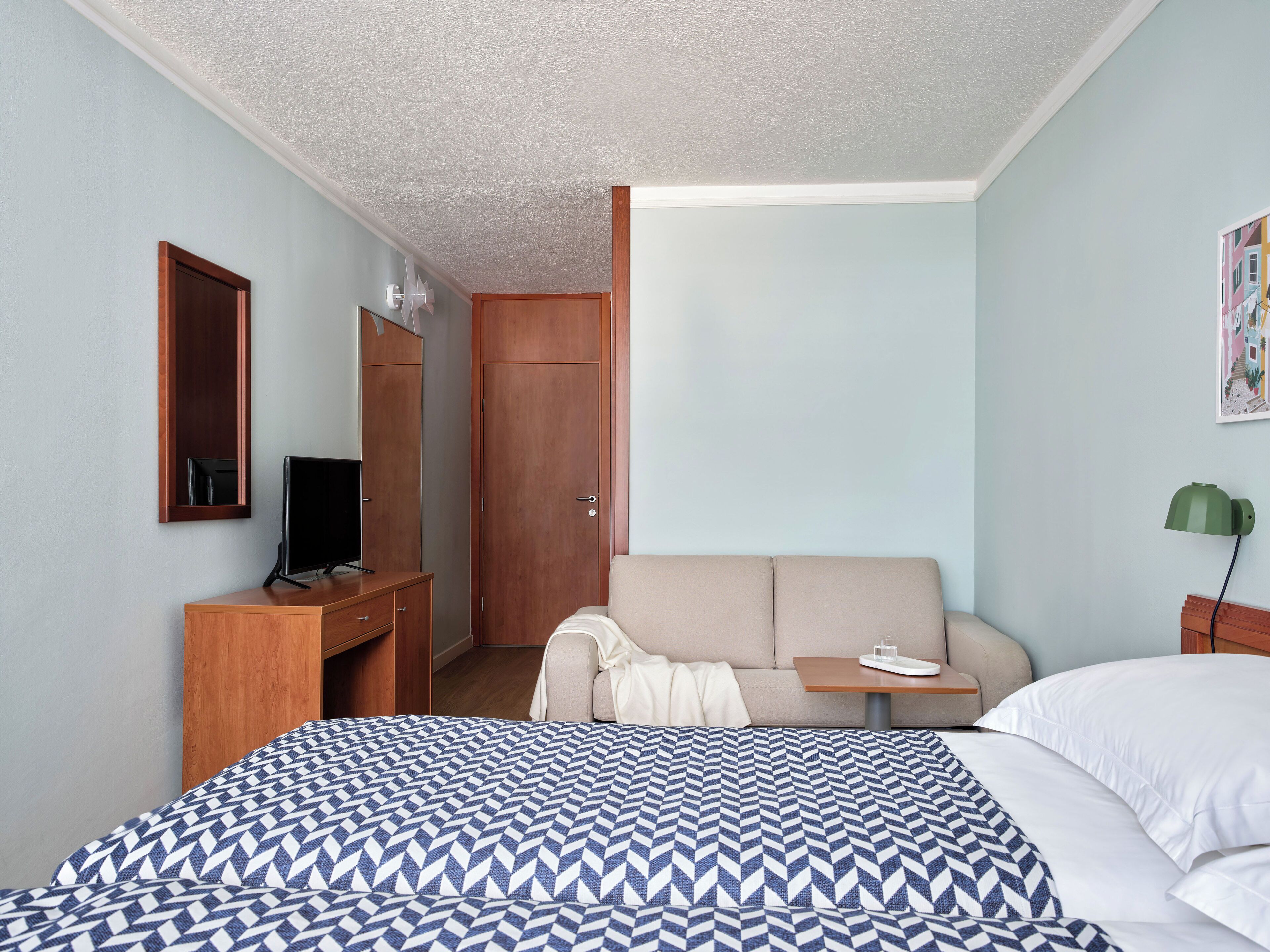 A hotel room with a bed, a desk with a computer, a sofa, a small table with a cup, and a door.