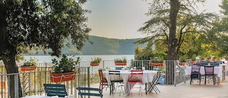 An outdoor dining area with tables and chairs, overlooking a body of water and hills.