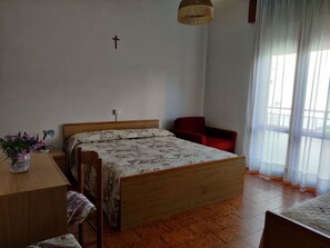 Een slaapkamer met een houten bed, een bureau met een lamp, een stoel en een kruis aan de muur.