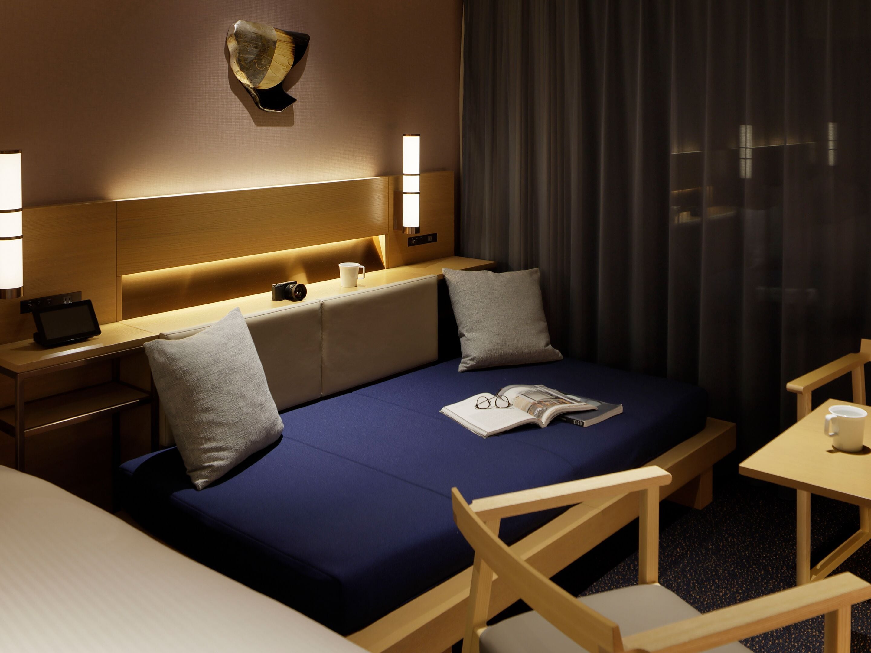 A hotel room with a bed, bedside tables, a lamp, a wall-mounted artwork, and a window with curtains.