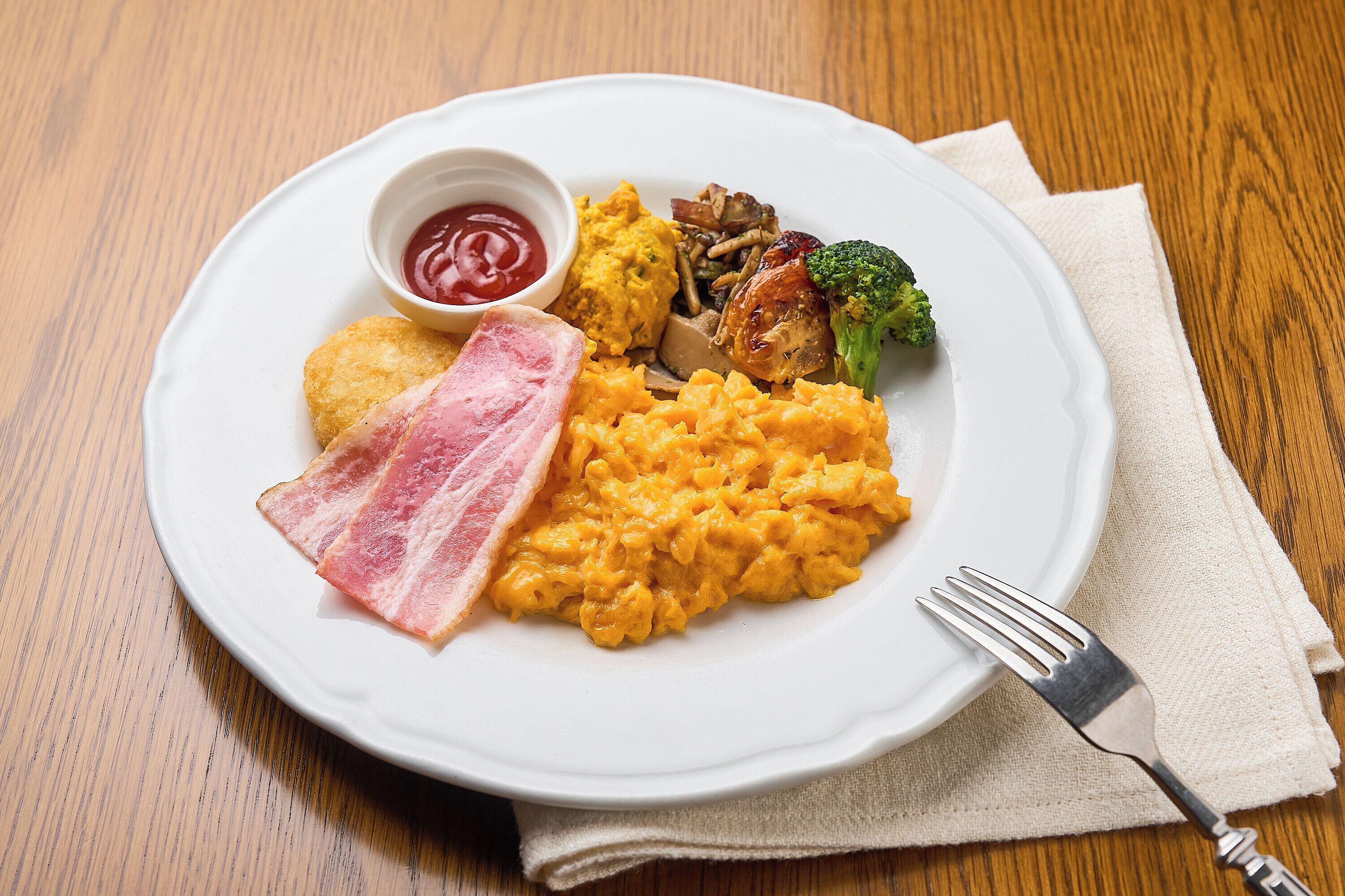 Um prato de comida com ovos mexidos, bacon, purê de batatas e acompanhamento de legumes.