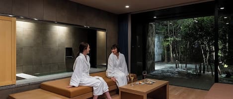 A modern spa room with a wooden bench, a small table, and a large window offering a view of a forested area.