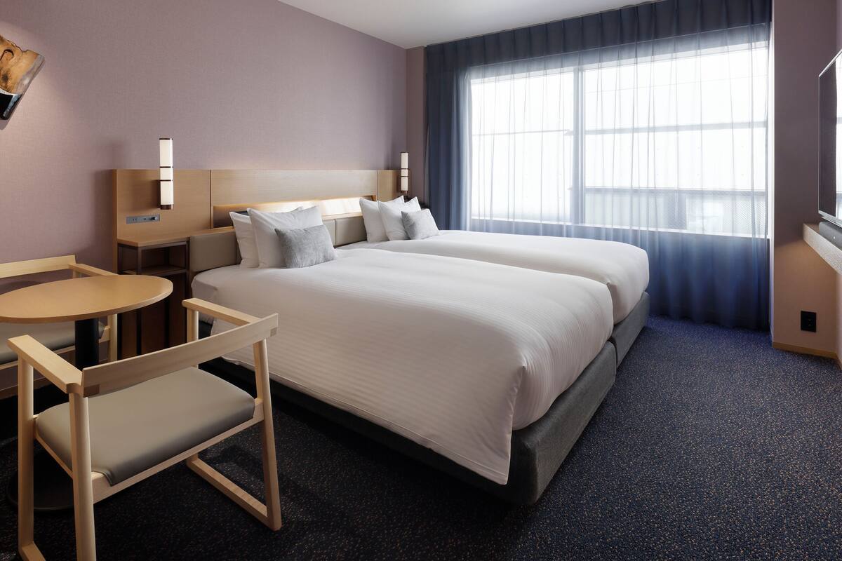 Ett hotellrum med två sängar, ett litet bord, en stol, en TV och ett stort fönster med gardiner.