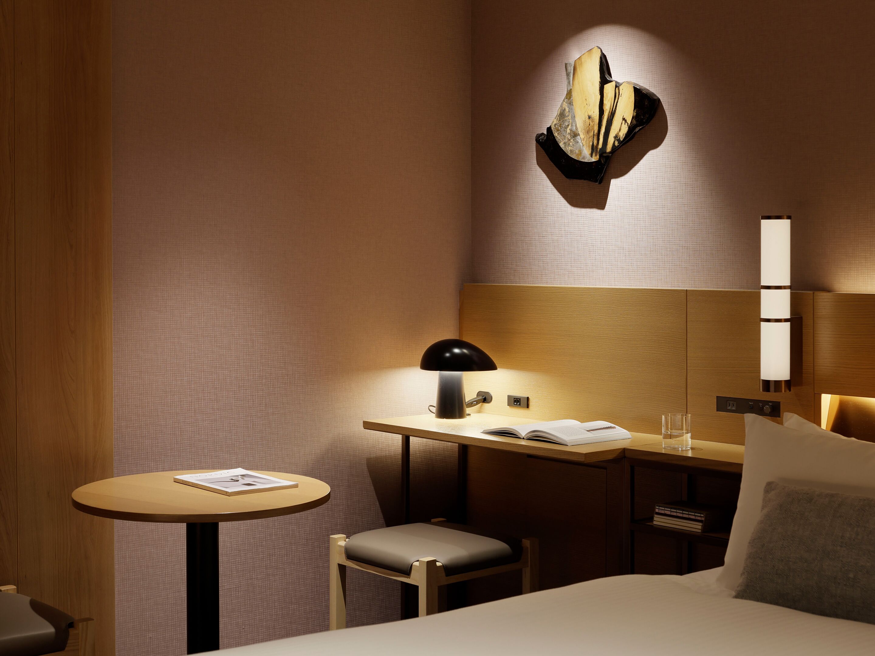 A hotel room with a bed, a desk with a lamp, a chair, a small round table with a book, and a wall-mounted light fixture.