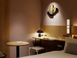 A hotel room with a bed, a desk with a lamp, a chair, a small round table with a book, and a wall-mounted light fixture.