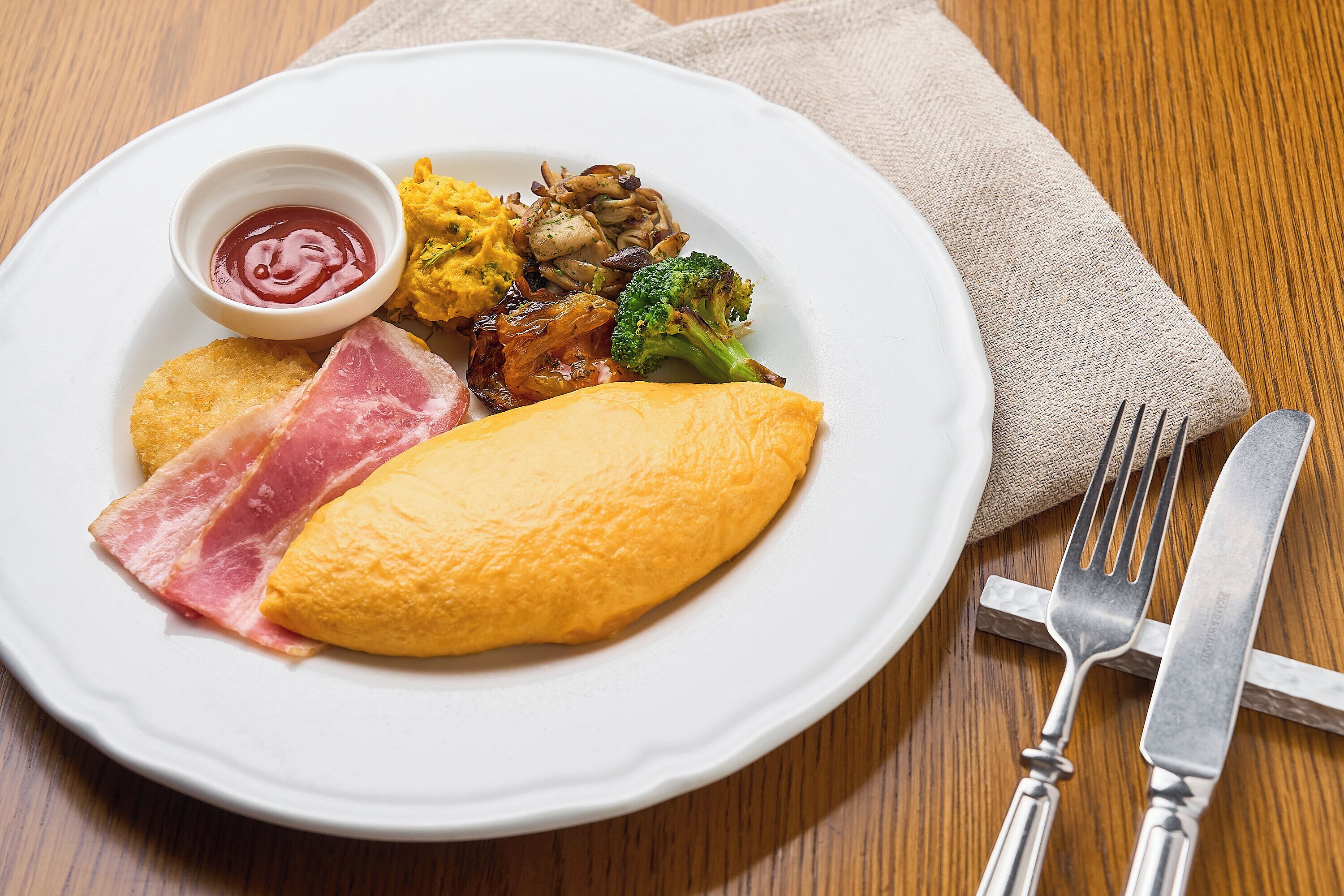Une assiette contenant divers aliments, notamment des œufs, du jambon, des champignons et des brocolis, accompagnée d’un petit bol de ketchup.
