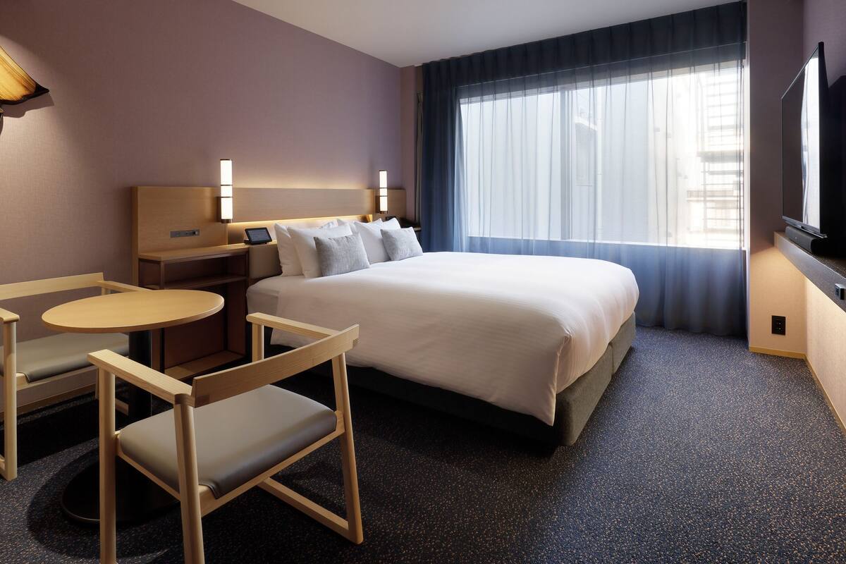 Ett hotellrum med en säng, sängbord, ett skrivbord, en stol, en TV och ett stort fönster.