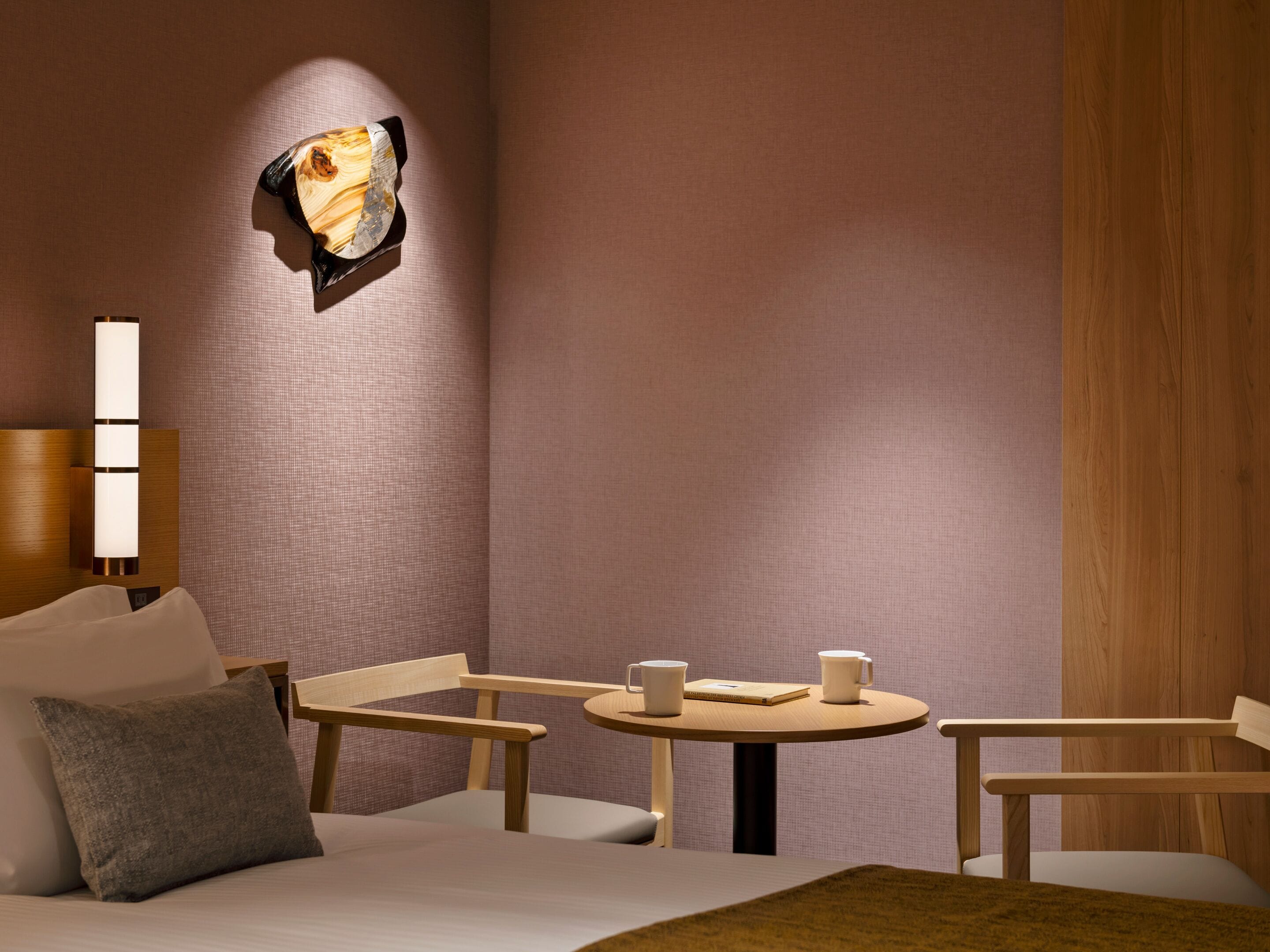 A modern hotel room with a bed, two chairs, a small table, and a wall-mounted light fixture.