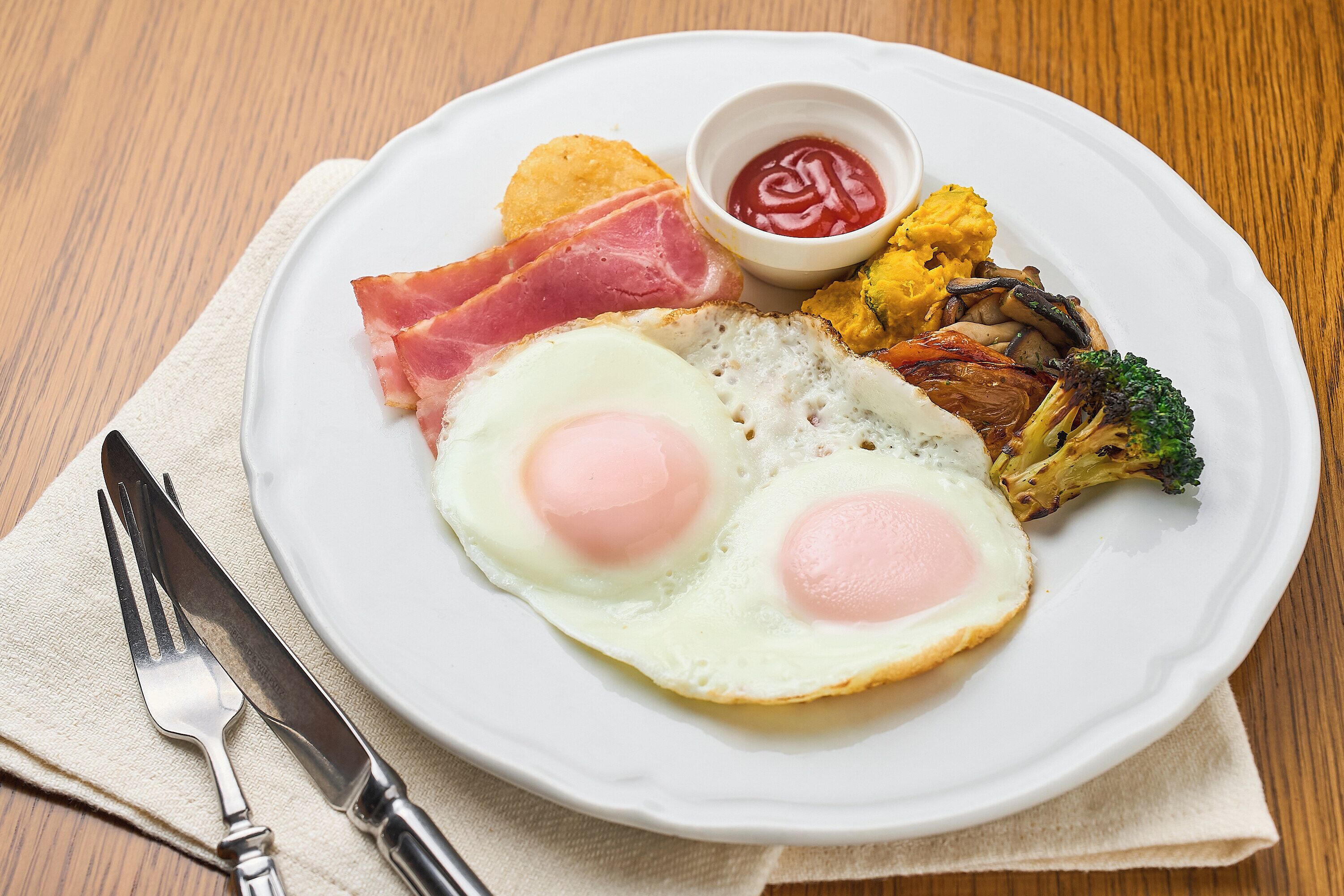 A plate with eggs, ham, toast, and vegetables on a wooden table.