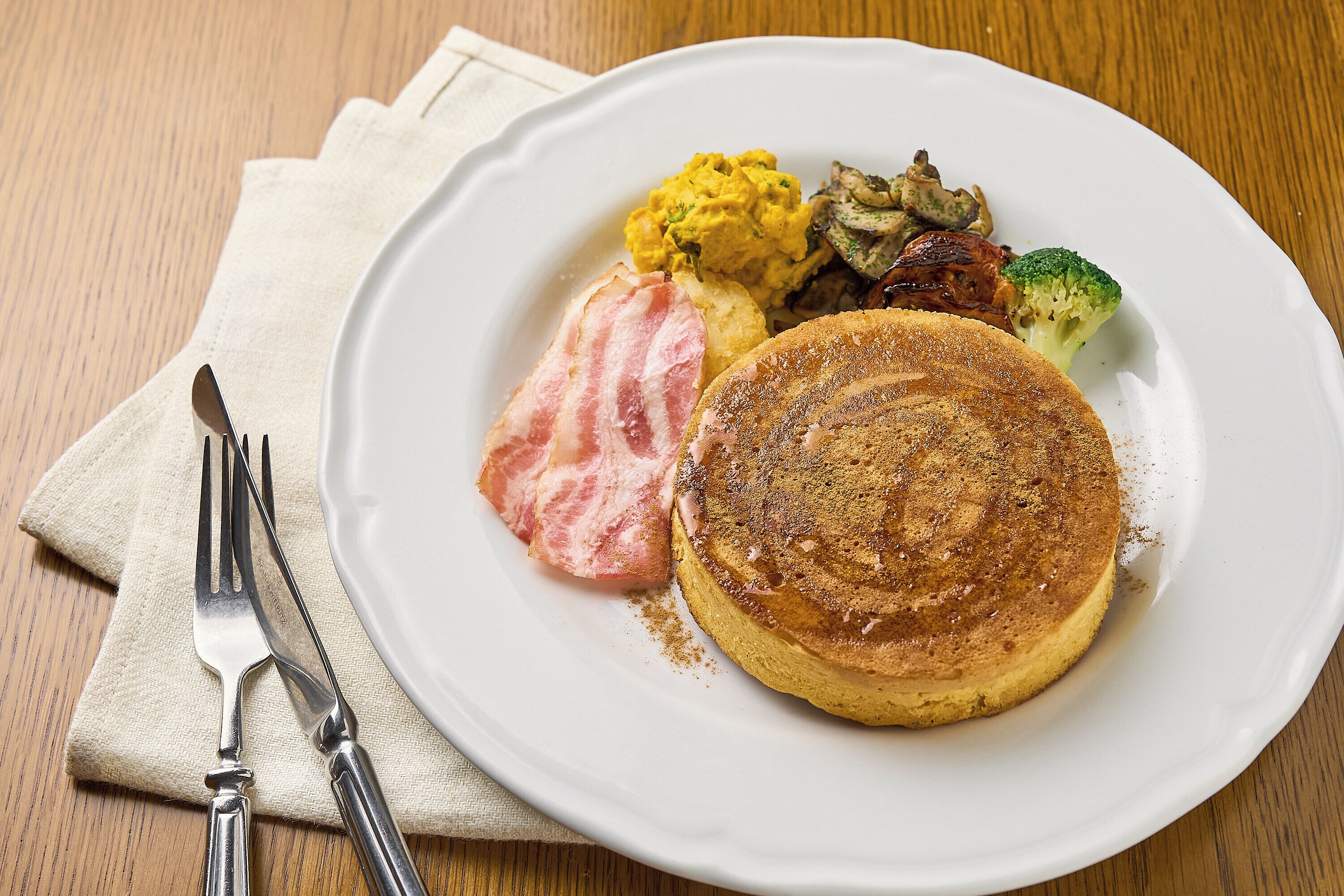 Een bord met pannenkoeken, spek, roerei en groenten op een houten tafel.