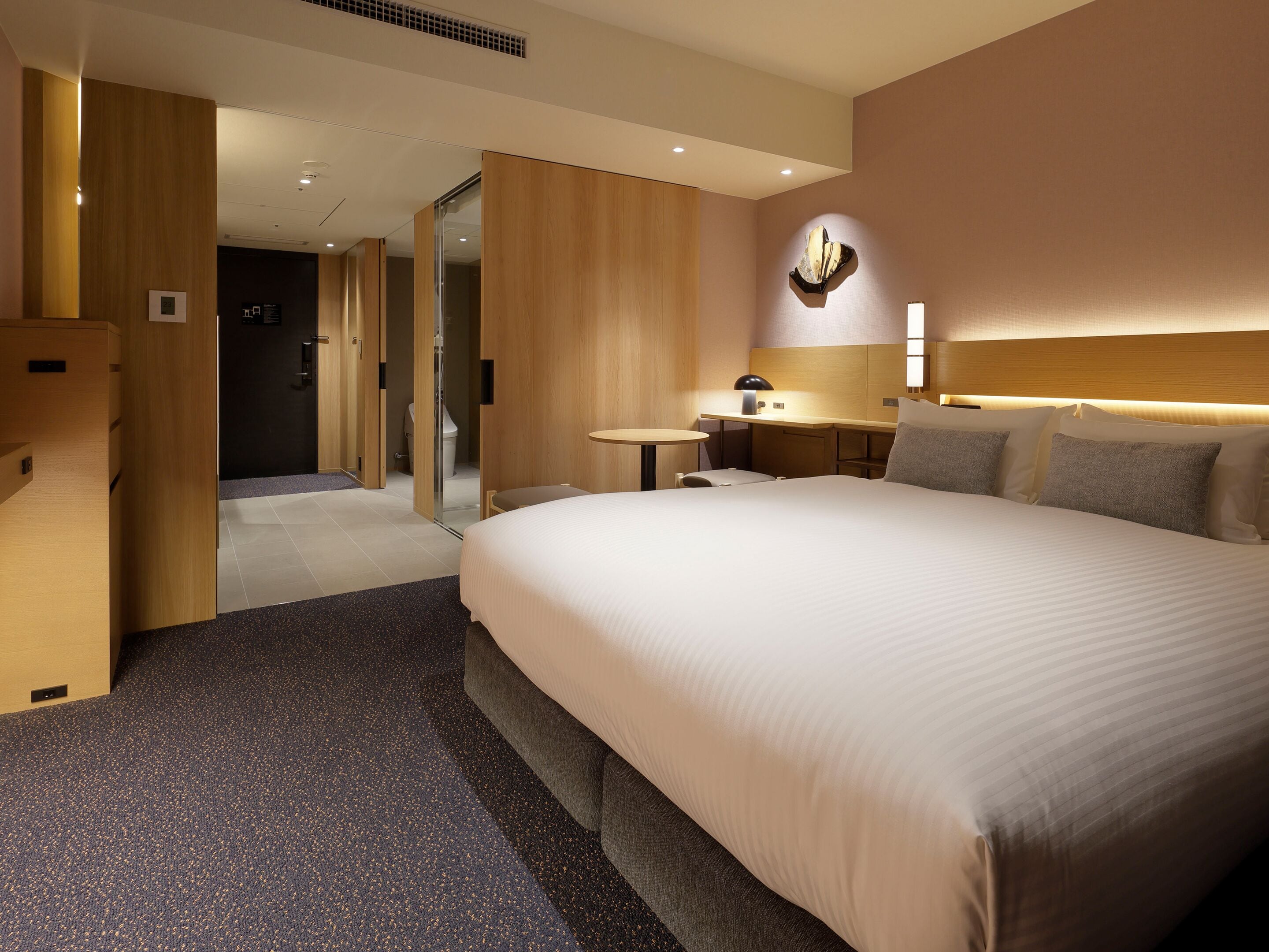 Habitación de hotel con una cama grande, un escritorio, una silla, una mesita y un baño visible a través de una puerta abierta.