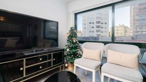 Una sala de estar moderna con un televisor de pantalla plana grande, dos sillones blancos, un pequeño árbol de Navidad y un ventanal con vistas a la ciudad.