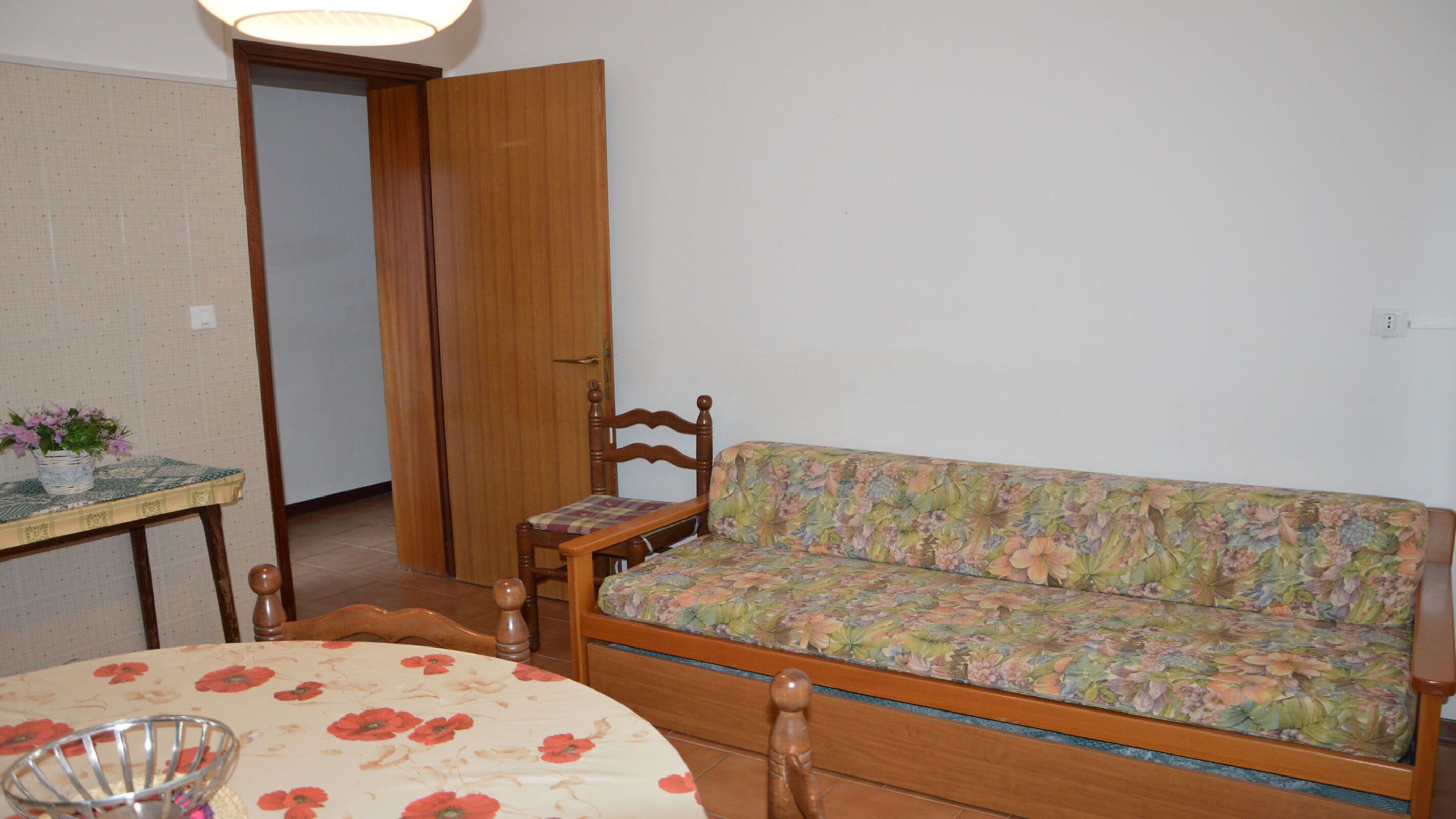 A room with a wooden sofa, a wooden chair, a small table with a floral tablecloth, and a potted plant.