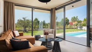 A modern living room with a sofa, a coffee table, and a chair, with a large sliding glass door leading to a pool and outdoor area.