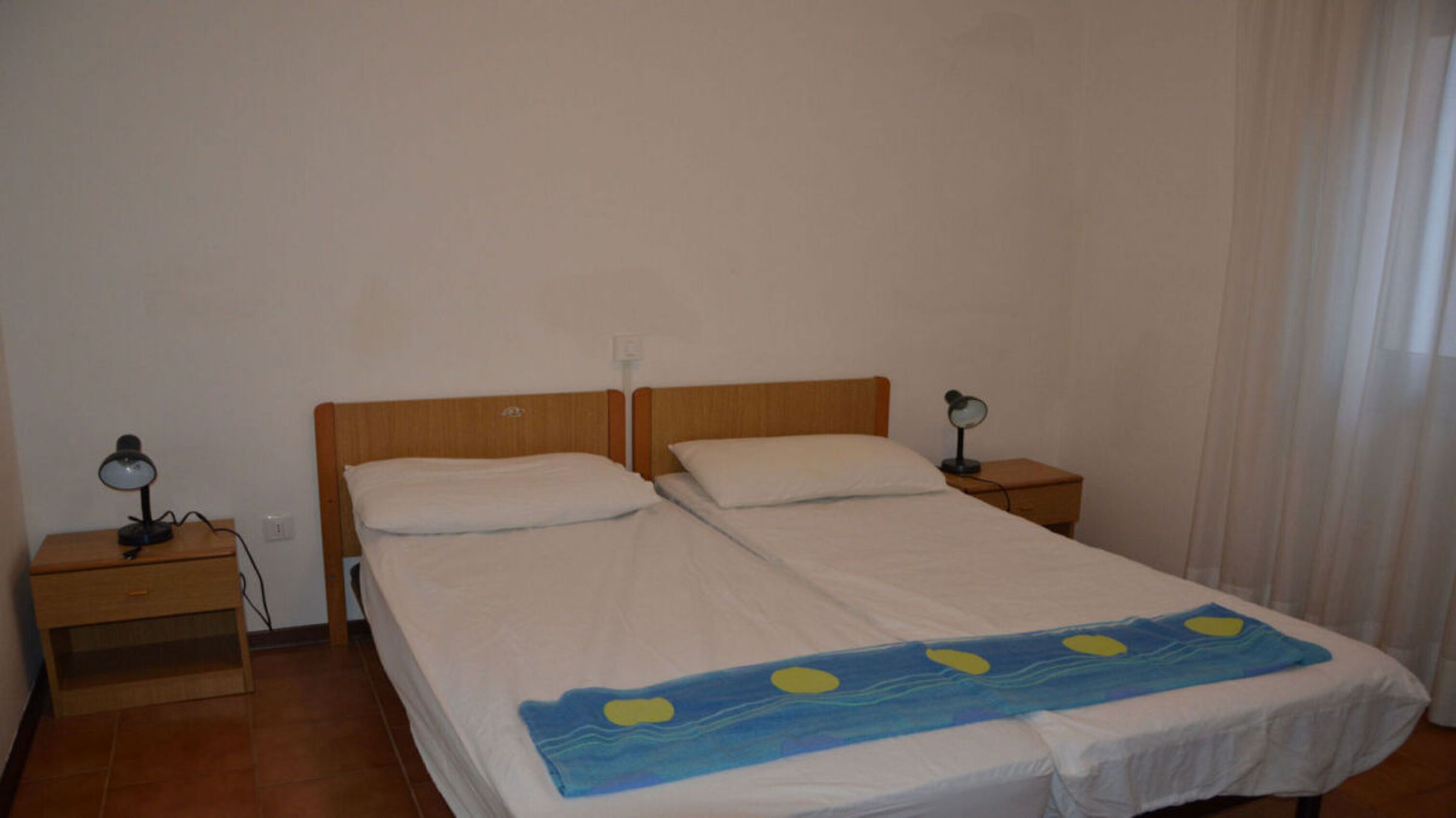 Habitación individual con una cama de una sola plaza, cabecera de madera, dos mesitas de noche con lámparas y una colcha con diseño azul y amarillo.