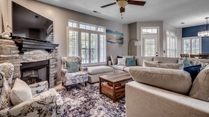 A living room with a stone fireplace, a ceiling fan, and a large window with blinds. - Shore Nuff: Surfside Beach. 1 blk to Beach, Private Pool, Golf Cart (Surfside Beach)