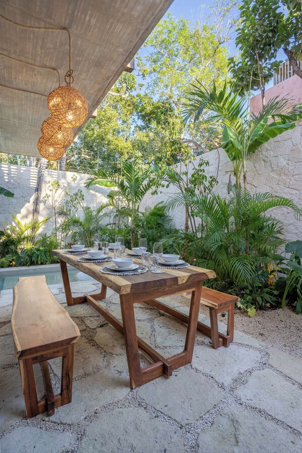 An outdoor dining area with a wooden table set for a meal, surrounded by lush greenery and a stone wall.