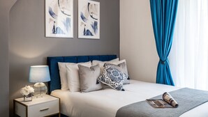 A neatly arranged bedroom with a bed, bedside table, lamp, and two framed abstract paintings. - Charming 4-Bedroom Apt Minutes from Borough St (Greater London)