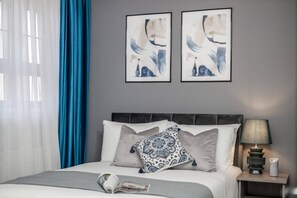 Een net opgemaakt bed met een grijs-wit kleurenschema, een gestreept kussen en een nachtkastje met een lamp en een kaars.