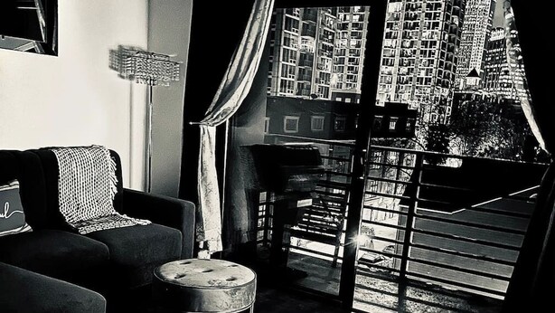 Een zwart-wit interieur met een bank, een kleine ronde tafel, een vloerlamp en een stadsgezicht te zien door een raam met jaloezieën.