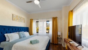 Een slaapkamer met een gebobbelde hoofdbord, een bed met wit en blauw beddengoed, een houten bureau met een televisie, en een raam met gordijnen.