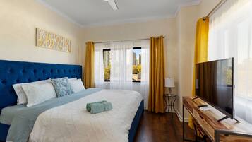 A bedroom with a tufted headboard, a bed with white and blue bedding, a wooden desk with a television, and a window with curtains.
