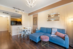 Una sala de estar moderna con un sofá azul, un otoman redondo, una mesa de comedor con sillas y una cocina al fondo.