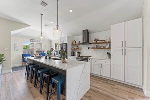 A modern kitchen with white cabinets, a marble island, and bar stools. - Stylish Home with Hot Tub & Gaming Garage (Spring)