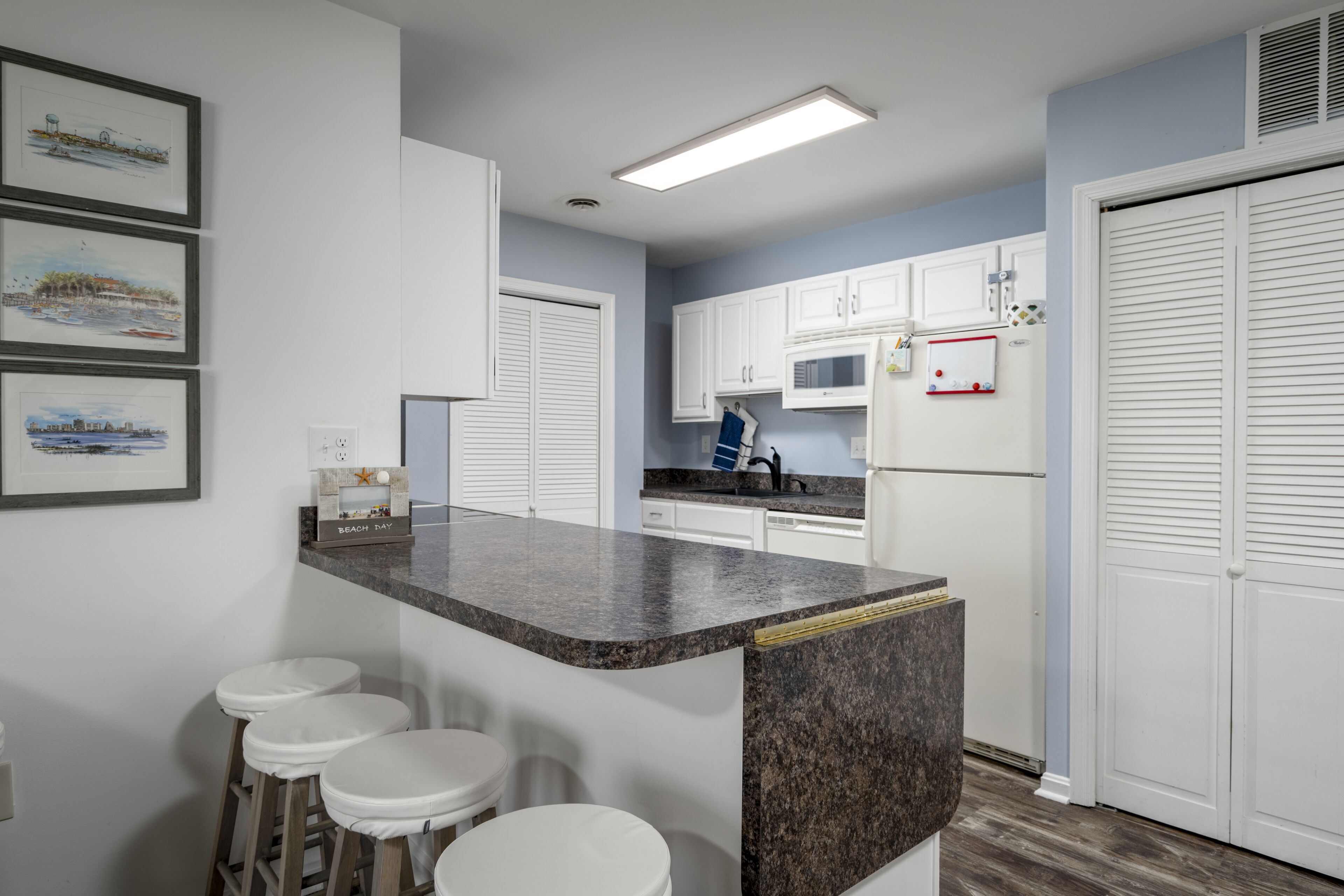 A kitchen with white cabinets, a granite countertop, and a microwave. There are three white stools and a small island with stools.