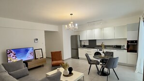 Een moderne woonkamer met een bank, een salontafel en een televisie. Er is een eethoek met stoelen en een tafel. De keuken is zichtbaar, met witte kastjes en apparatuur.