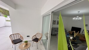 Een moderne woonruimte met een glazen schuifdeur naar een balkon, ingericht met rieten stoelen en een kleine ronde tafel. Binnen is er een bank, een salontafel en een kroonluchter.