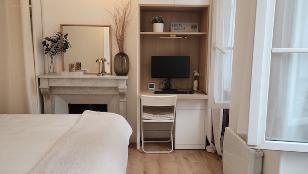 A modern bedroom with a large bed, a desk with a computer, a small shelf with decorative items, and a window. - Appartement Montparnasse - rue de la Gaieté (Paris)