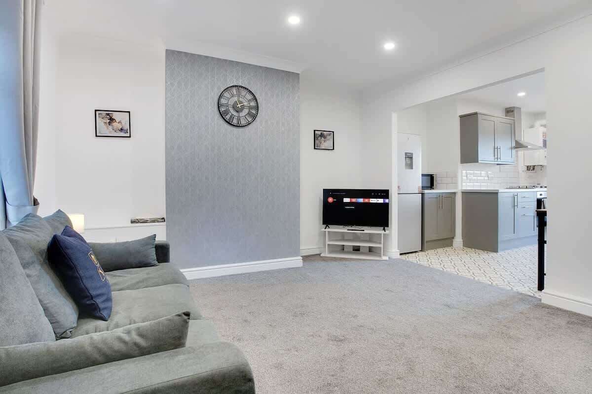 Een moderne woonkamer met een grijze bank, een flatscreen televisie en een keuken op de achtergrond.