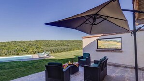 Eine Terrassengaststätte mit Korbmöbeln, einem großen Sonnenschirm und Blick auf ein Schwimmbecken und eine grüne Landschaft.