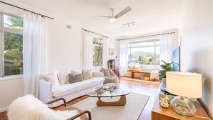 A spacious living room with a white sofa, a glass coffee table, and a view of greenery outside.
