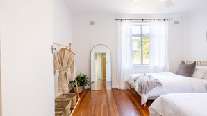 A bedroom with a wooden floor, a bed with white bedding, a bedside table, a mirror, and a window with curtains.