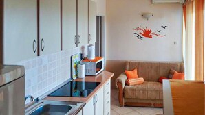 Eine kleine Küche mit Mikrowelle, Wasserkocher und einer kleinen Arbeitsfläche. Im Wohnbereich steht ein beiges Sofa mit orangefarbenen Kissen, und an der Wand ist eine Vogelsticker zu sehen.