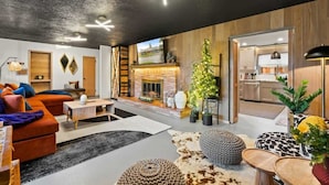 A modern living room with a fireplace, a kitchen in the background, and a dining area with a table and chairs. - Chic Newberg Oasis: Mid-Century, Hot Tub, Fire Pits, Luxury Amenities (Newberg)