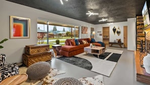 A modern living room with a large window, a brown sofa, a coffee table, and decorative items. - Chic Newberg Oasis: Mid-Century, Hot Tub, Fire Pits, Luxury Amenities (Newberg)