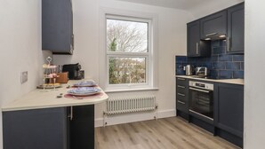 A modern kitchen with dark cabinets, a white countertop, and a window with a view of trees. - Sally (Whitstable)