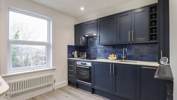 A modern kitchen with dark cabinets, a white countertop, and a window providing natural light. - Sally (Whitstable)