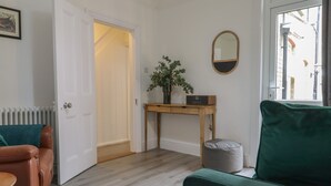 Ein Zimmer mit Holzboden, einem grünen Sofa, einem braunen Ledersessel, einem Holztisch mit einem Radio, einem Spiegel und einer Tür, die in einen anderen Raum führt.