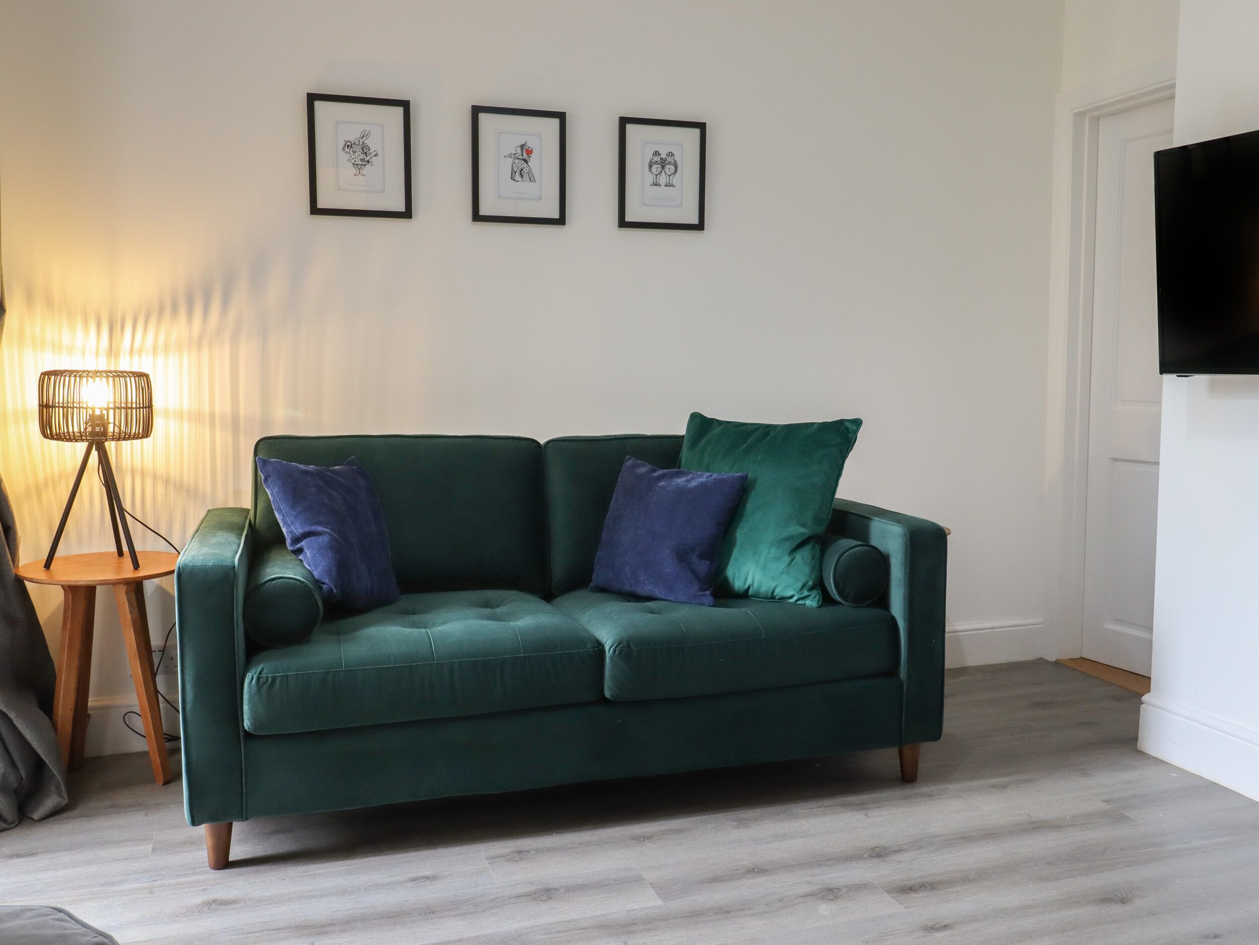 A green sofa with blue and green cushions, a floor lamp, and three framed pictures on the wall.