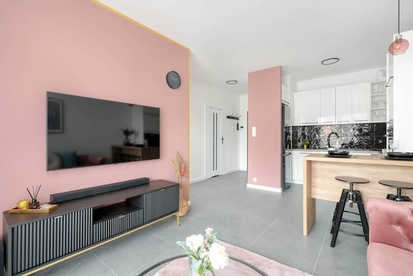 A modern living room with a large flat-screen TV, a pink accent wall, and a kitchen area with a wooden countertop and stools.